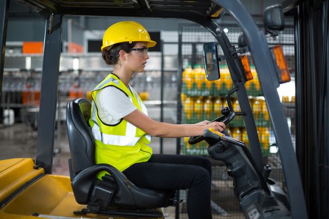 Female factory worker driving forklift in warehouse, wearing safety gear including hard hat and high visibility vest. Ideal for illustrating industrial work environments, logistics, transportation, and occupational safety. Useful for articles on manufacturing, warehouse operations, and workplace safety protocols.