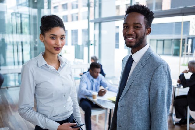 Two business colleagues are standing and smiling in a modern office environment. They are dressed in formal attire, suggesting a professional setting. This image can be used for promoting corporate culture, teamwork, diversity in the workplace, or business success stories.