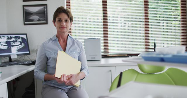 Female dentist is seen sitting in a modern dental office holding patient records. This scene portrays a professional medical environment suitable for healthcare, dental care services, and professional medical consultation advertisments. The dentist's friendly demeanor is appealing for promoting dental practices, healthcare facilities, and patient-doctor interactions.