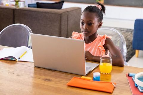 Young girl studying on laptop at home desk with notebook, school supplies and orange juice