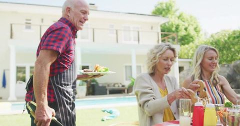 Senior man serving family poolside barbecue holding plate and tongs