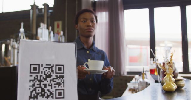 This image shows a barista holding a cup of coffee, standing behind a cafe counter with a QR code sign. The QR code implies modern and contactless technological solutions, which are gaining popularity in the service industry. This scene portrays a warm and inviting feeling of a small business. Ideal for use in promotions for cafes, articles about service innovation, or visual content related to hospitality and customer service.