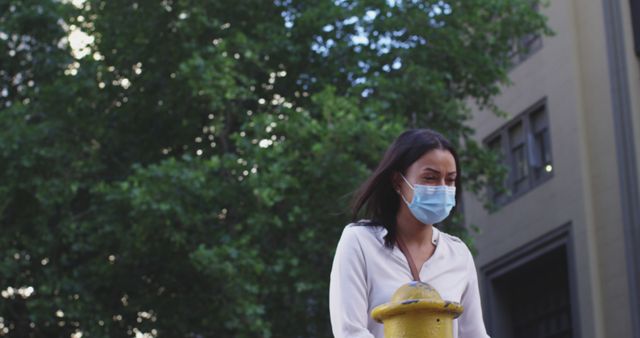 Young woman wearing a protective face mask while outside in an urban environment. Trees and buildings can be seen in the background. Suitable for content about health and safety, pandemic guidelines, and urban life.