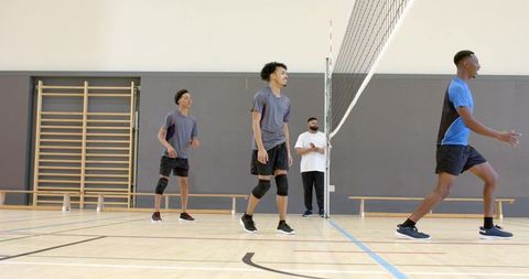 Diverse male volleyball team practicing drill on indoor court with coach observing