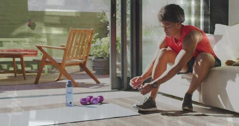 Tying running shoes before indoor workout by sliding glass door with mat and dumbbells