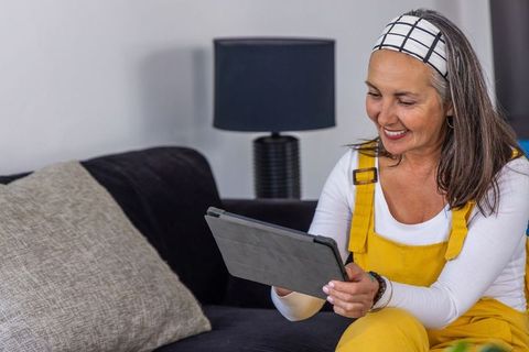 Senior woman smiling while using tablet on sofa wearing yellow overalls and headband