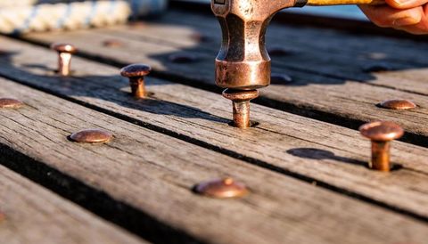 Claw hammer driving copper fastener into weathered wooden deck planks in warm sunlight