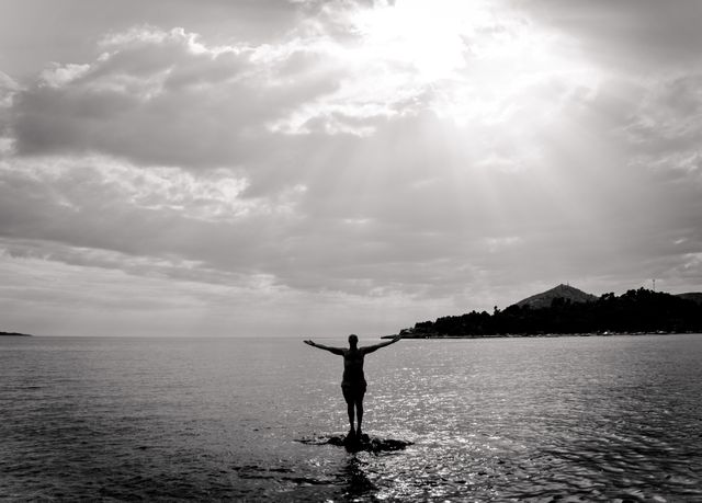 Person stands alone on shore with arms outstretched facing sunrise. Black and white theme enhances calm and serene mood. Useful for themes like solitude, nature's beauty, introspection, new beginnings, and peace. Ideal for wellness, meditation, and travel-related content.