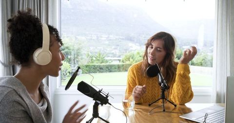 Female cohosts recording podcast at bright cozy home table with microphones and laptop