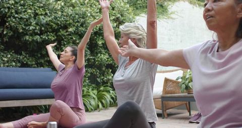Diverse women practicing seated yoga on brick patio with plants and wellness props