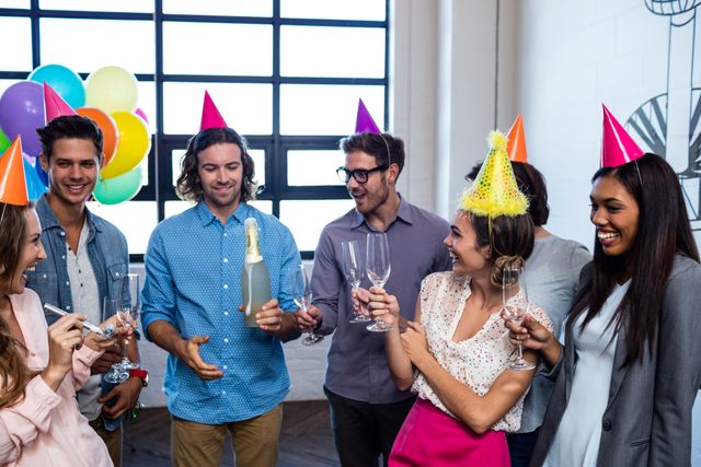 Coworkers gather around to celebrate a birthday in the office. One person is opening a bottle of champagne while others hold glasses and wear party hats. Balloons are visible in the background, adding to the festive atmosphere. This image is perfect for illustrating workplace culture, team bonding, and office celebrations.