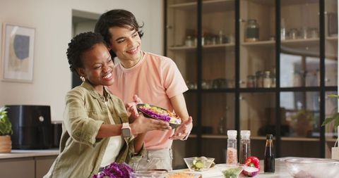 Diverse couple preparing colorful salad together in modern kitchen, smiling and sharing