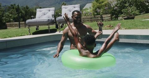 African american father and son playing in backyard pool with bright green float