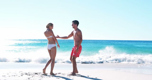 Young couple in swimwear playing on a sunny beach with ocean waves in the background. Ideal for ads related to beach vacations, summer products, travel destinations, leisure activities, and romantic getaways.