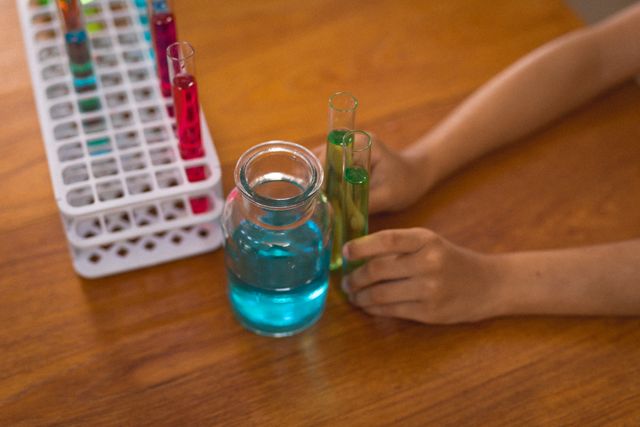 Hands of hispanic boy holding test tubes with chemical solutions by beaker and rack on table at home. unaltered, childhood, science experiment, discovery and learning concept.