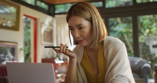 Asian woman sitting at table working from home talking on smartphone and using laptop. at home in isolation during quarantine lockdown.