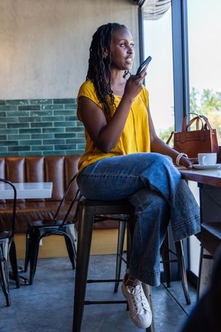 African american woman using smartphone at cafe counter wearing yellow blouse and jeans
