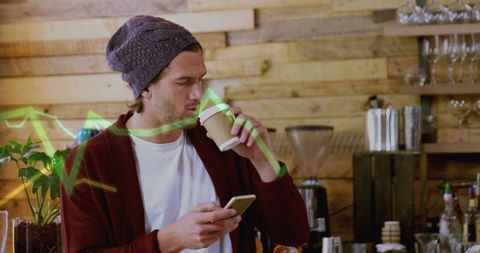 Man holding takeaway coffee checking smartphone with green stock chart overlay in wood cafe