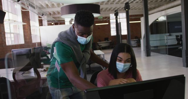 Diverse male and female business colleagues wearing face masks in discussion at work using computer. business in modern office during covid 19 coronavirus pandemic.