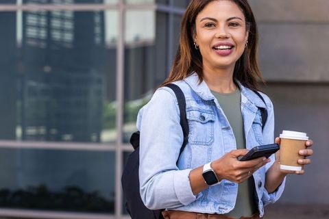 African american commuter smiling with smartphone and coffee outside glass office building