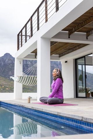 Senior woman meditating by pool on terrace wearing headset and purple activewear