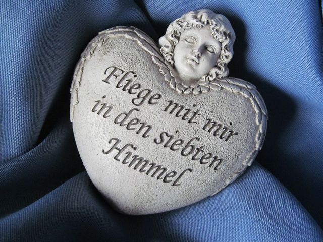 Heart-shaped stone with an angel figure and a German inscription on blue fabric conveying a sense of tranquility and heavenly peace. Ideal for use in topics related to inspiration, religious faith, remembrance, decorative arts, and sentiments of love and peace.