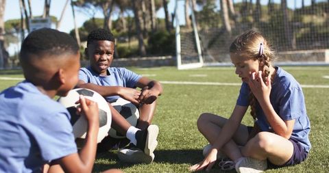 Diverse teenagers sitting on soccer turf sharing balls during sunlit team break