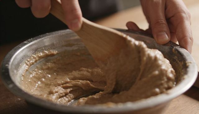 Hands stirring rustic whole-grain batter in metal bowl with wooden spoon sunlit kitchen