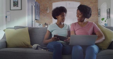 African american mother and teen daughter sharing tablet and laptop on cozy sofa