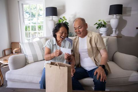 Asian couple opening surprise card and gift on sofa in sunlit modern living room