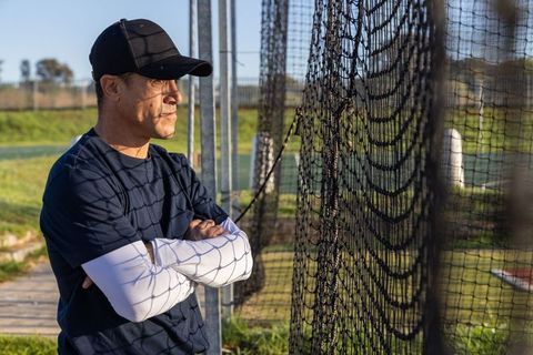 Mature coach observing batting cage during outdoor baseball training with net shadow