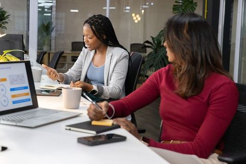 Diverse women collaborating on laptop charts and taking notes during office meeting