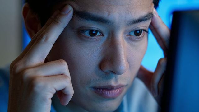 Young asian man pressing temples and focusing on glowing monitor, late night concentration