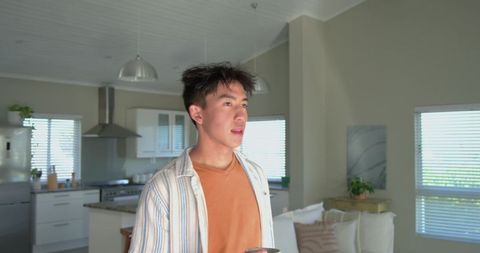 Asian young man standing in minimalist open-plan kitchen holding mug and looking right