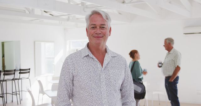 Mature man with white hair smiling at a social gathering, wearing a casual shirt. A woman and another man in background engaging in conversation. White, bright, airy indoor space. Perfect for use in contexts related to social events, community gatherings, casual meetings, or modern indoor venues.