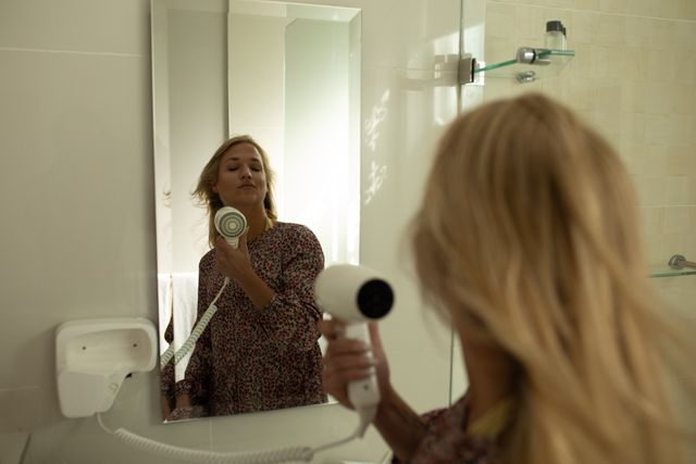 Young Caucasian woman drying her long, blonde hair with a hairdryer, reflected in a bathroom mirror. Ideal for use in travel, hospitality, self-care, and beauty-related content, showcasing relaxation and morning routines in an elegant hotel setting.