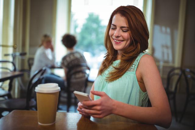 Young woman sitting at a table in a cafe, smiling while using her smartphone. A coffee cup is on the table. Ideal for use in lifestyle blogs, social media posts, advertisements for cafes or coffee shops, and articles about technology and communication.