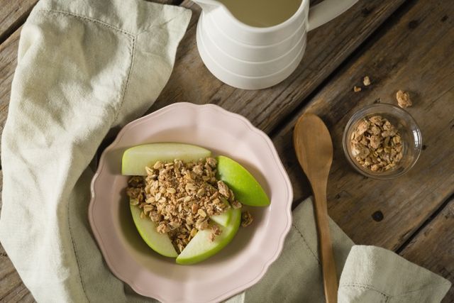 Overhead view of a healthy breakfast featuring granola and green apple slices on a pink plate. The rustic wooden table and natural light create a warm, inviting atmosphere. Ideal for use in food blogs, healthy eating promotions, breakfast menus, and lifestyle magazines.