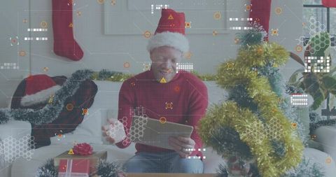 Smiling man wearing Santa hat using tablet in living room with Christmas tree and gifts