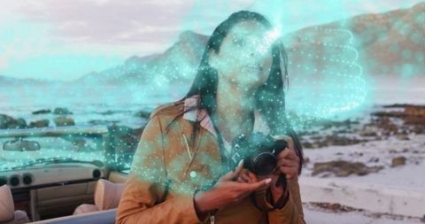 Indian woman holding DSLR on rocky coastal beach with convertible and turquoise hologram