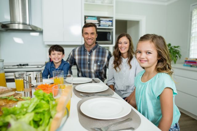 Family sitting together at dining table in kitchen, smiling at camera. Fresh salads, drinks, and tableware visible. Ideal for concepts of family bonding, indoor activities, healthy eating, and home life.