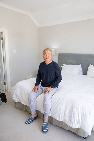 Mature chinese man sitting on bed edge in bright minimalist bedroom wearing slippers