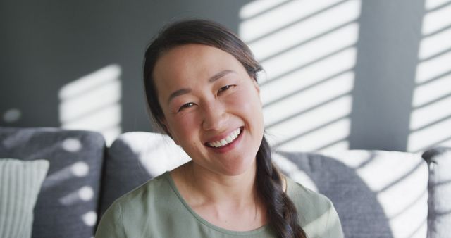 Asian woman sitting comfortably on a gray sofa, smiling warmly. Natural light from window blinds creates patterns on the wall. Perfect for themes of relaxation, comfort, home lifestyle, and happiness, suitable for advertisements, social media posts, and blog articles about home living and mental well-being.