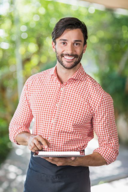 Smiling male waiter using digital tablet in an outdoor cafe. Ideal for illustrating modern restaurant technology, customer service, and hospitality industry. Perfect for use in articles, advertisements, and promotional materials related to dining, service, and technology integration in restaurants.