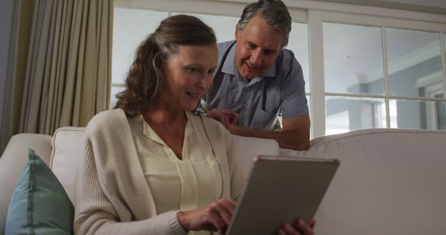 Happy senior caucasian couple in living room using tablet and talking. retirement lifestyle at home.