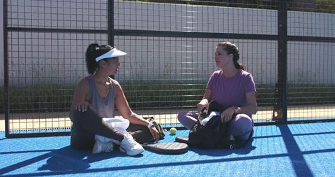 Two women chatting on blue padel court resting after match sharing gear and laughing