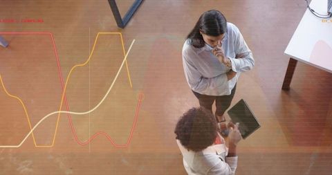 Two businesswomen analyzing tablet with data visualizations from overhead in modern office