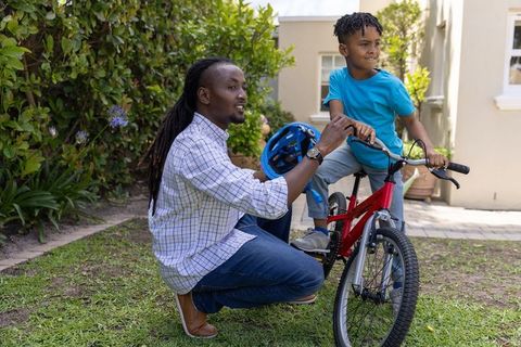 African american father kneeling teaching son wearing helmet riding bike in sunny backyard