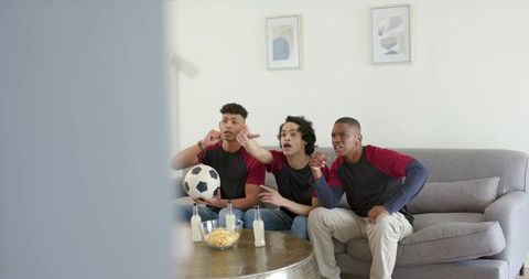 Friends reacting to soccer game on couch, holding soccer ball and sharing snacks