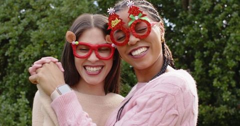 Female friends wearing festive red glitter glasses holding hands smiling in garden